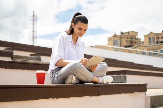 Heureuses jeunes femmes tenant une tablette avec une tasse de papier café profitant d'une journée ensoleillée assise dans les escaliers de la ville