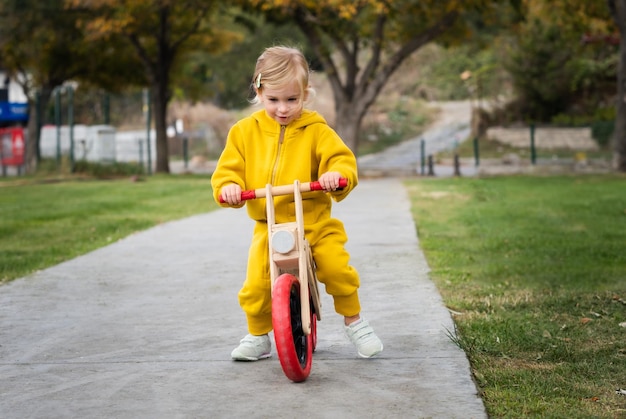 Heureux actif assez mignon caucasien blond bébé fille enfant enfant en bas âge souriant enfant d'environ 2 ans portant des cavaliers jaune vif apprenant à faire du vélo d'équilibre dans le parc d'été à l'extérieur