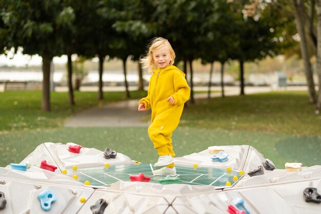 Photo heureux actif assez mignon caucasien blond bébé fille enfant enfant en bas âge souriant enfant d'environ 2 ans portant des pulls jaune vif dansant sur l'aire de jeux dans le parc d'été à l'extérieur