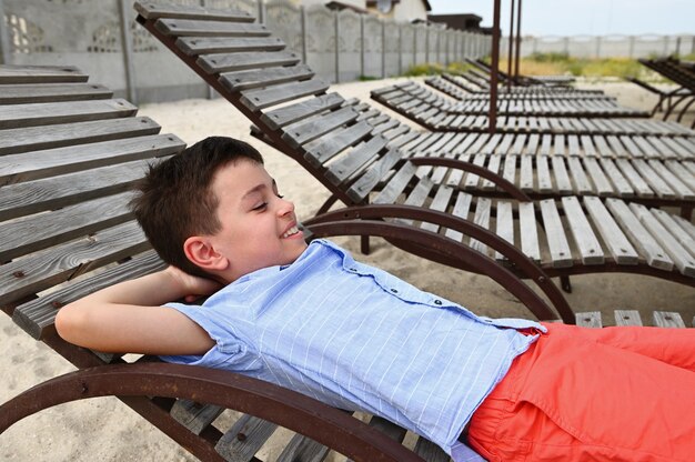 Heureux adolescent allongé sur une chaise longue en bois. Bel enfant reposant sur la chaise longue pendant les vacances d'été à la campagne. Enfants en vacances