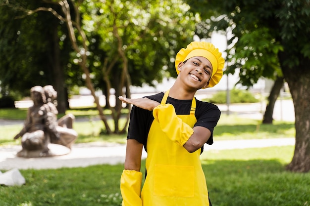 Heureux adolescent noir africain cuisinier en toque de chefs et uniforme de tablier jaune souriant en plein air Publicité créative pour café ou restaurant