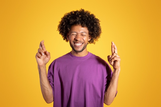 Heureux adulte afro-américain bouclé homme en t-shirt violet avec les yeux fermés les doigts croisés fait un vœu