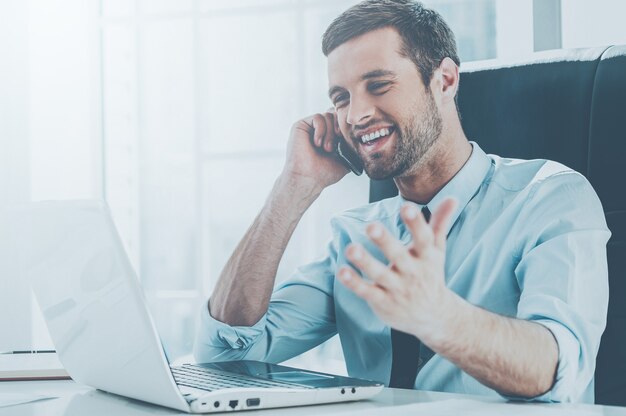 Heureux avec les affaires en cours. Heureux jeune homme en tenue de soirée regardant un écran d'ordinateur et parlant au téléphone portable alors qu'il était assis sur son lieu de travail