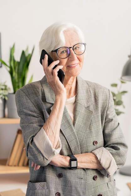 Heureux âgé de femme aux cheveux blancs dans des lunettes et des vêtements de cérémonie debout tout en parlant sur smartphone