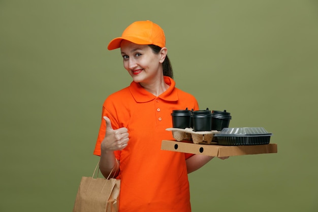 heureux d'âge moyen livraison femme portant l'uniforme et la casquette tenant un paquet de pizza plateau d'oeufs tasses à café en plastique récipient de nourriture sac en papier regardant la caméra montrant le pouce vers le haut