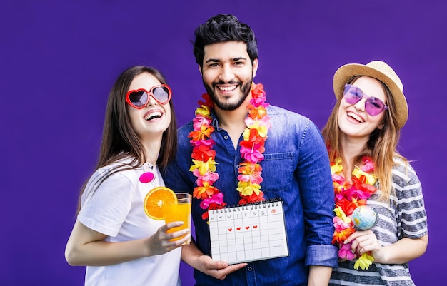 Heureux amis souriants deux filles et un homme barbu tiennent un calendrier de voyage de vacances avant le fond violet