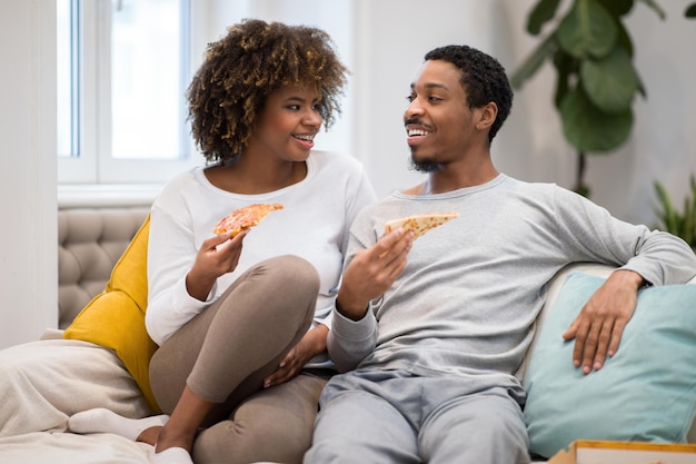 Heureux amoureux noirs assis sur un canapé en train de manger de la pizza