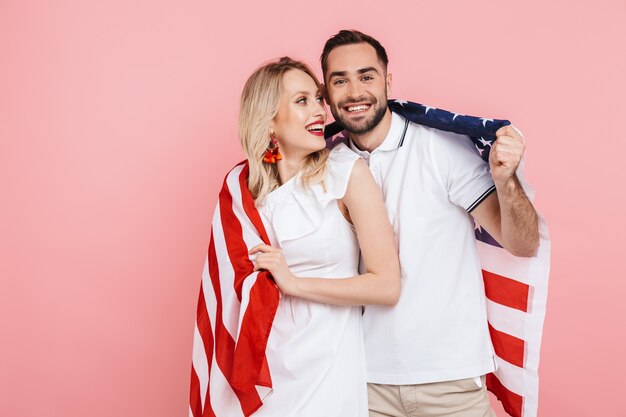 Heureux beau couple debout isolé sur rose, portant le drapeau américain, célébrant