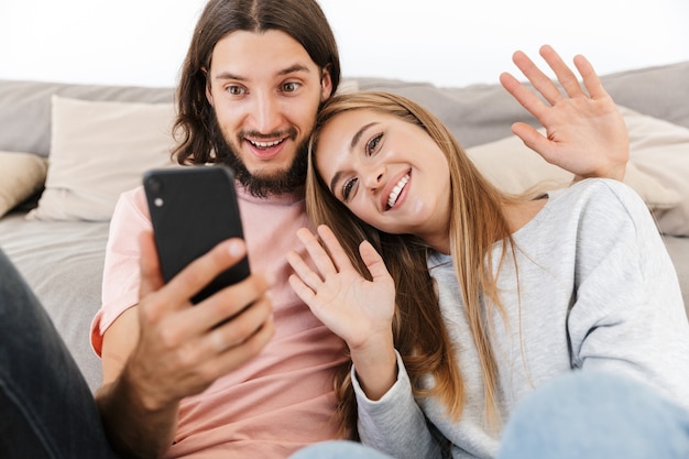 Heureux beau couple se détendre sur un canapé à la maison, passer un appel vidéo tout en tenant un téléphone portable, agitant les mains