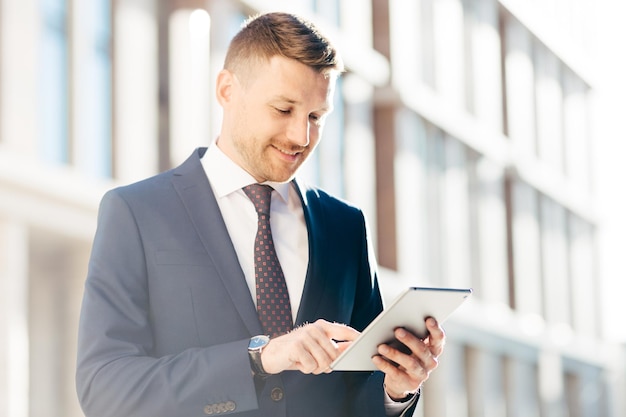 Heureux beau directeur exécutif masculin en costume élégant formel vérifie le message de revenu via une tablette numérique a un regard ravi pose en plein air Concept de réussite et de professionnalisme des gens d'affaires