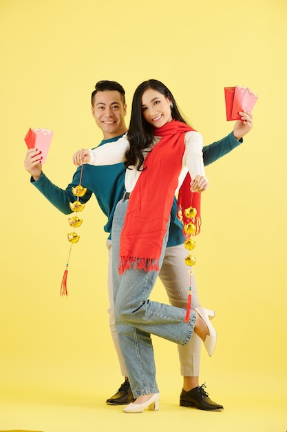 Heureux beau jeune couple asiatique en vêtements d'hiver montrant des enveloppes d'argent chanceux et des guirlandes de sycee ou de lingots d'or chinois