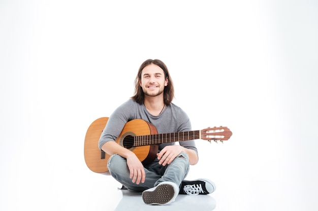 Heureux beau jeune homme aux cheveux longs assis sur le sol avec guitare sur fond blanc