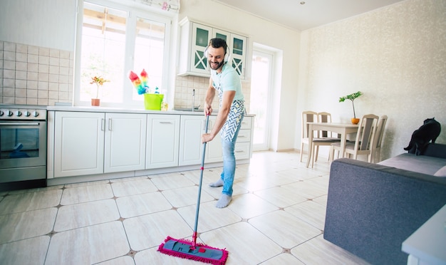 Heureux beau jeune homme barbe nettoie le sol dans la cuisine domestique et amusez-vous.