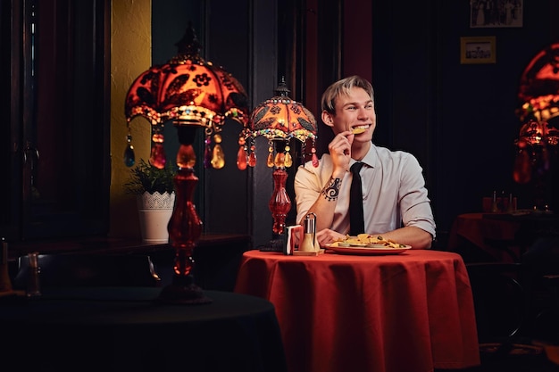 Heureux beau jeune homme en chemise blanche et cravate dégustant des nachos au restaurant mexicain.