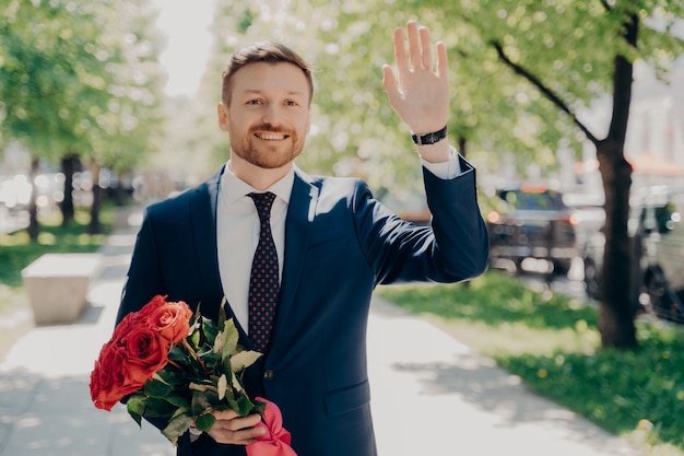 Heureux beau jeune homme en costume bleu tenant un bouquet frais de roses rouges et saluant la femme bien-aimée avec des fleurs tout en ayant un rendez-vous romantique dans le parc de la ville, souriant et se sentant heureux