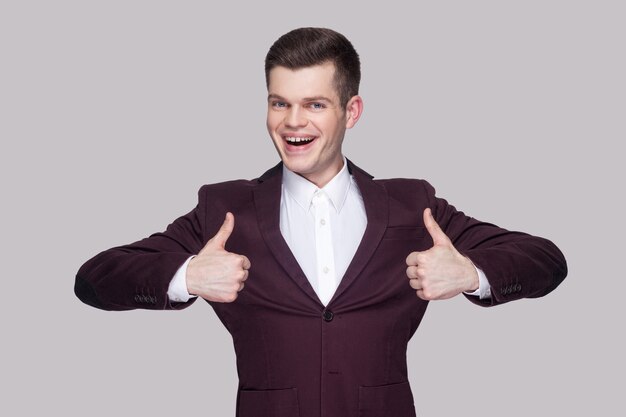 Heureux beau jeune homme satisfait en costume violet et chemise blanche, debout, bras levés et regardant la caméra avec les pouces vers le haut et le sourire à pleines dents. tourné en studio intérieur, isolé sur fond gris.