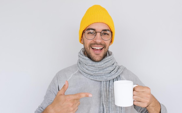Heureux beau mâle barbu avec une tasse de thé blanche portant un chapeau jaune et un pull gris avec une écharpe en automne ou en hiver Joyeux homme souriant annonçant quelque chose sur une tasse blanche Gens de médecine en bonne santé