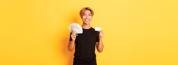 Heureux beau mec asiatique montrant de l'argent et une carte de crédit souriant heureux debout fond jaune