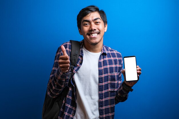 Heureux bel étudiant asiatique portant une chemise de flanelle tenant et pointant sur un écran de téléphone intelligent vierge sur fond bleu. Le gars montre une expression choquée et stupéfaite.