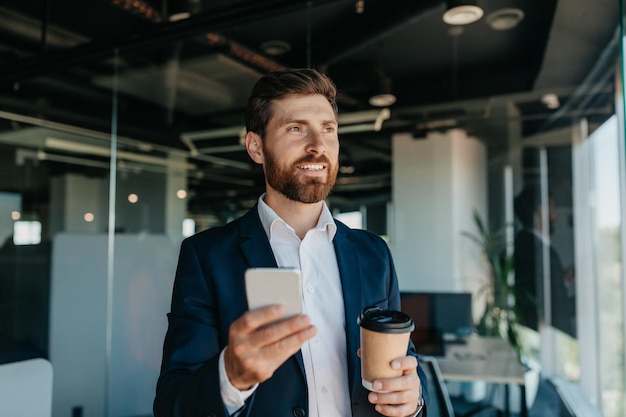 Heureux bel homme d'affaires en costume avec une tasse de café à l'aide d'un smartphone ayant une pause dans l'espace de copie intérieur du bureau