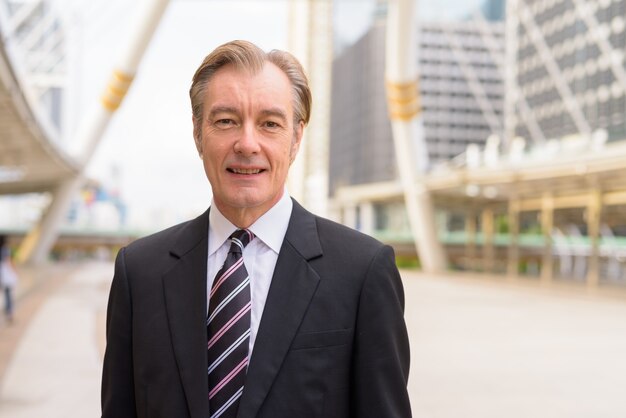 Heureux bel homme d'affaires mature en costume souriant au pont de skywalk dans la ville