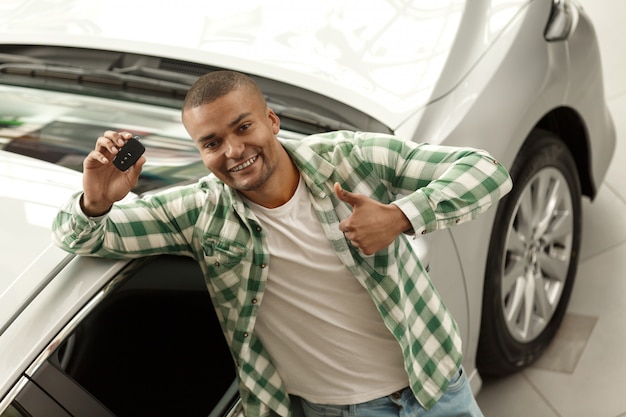 Photo heureux bel homme africain montrant le pouce en l'air tenant les clés de la voiture chez le concessionnaire.