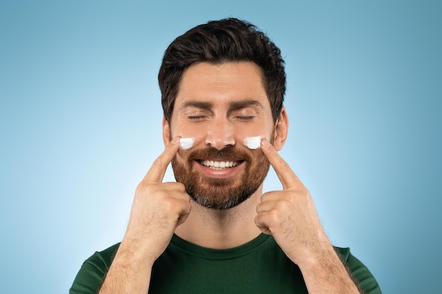 Heureux bel homme barbu appliquant de la crème pour le visage sur les joues avec les yeux fermés posant sur fond de studio bleu