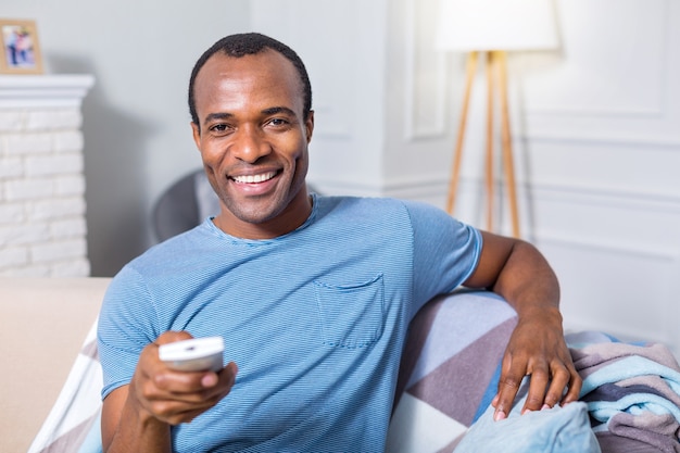 Heureux bel homme positif assis devant l'écran du téléviseur et tenant la télécommande tout en se reposant sur le canapé