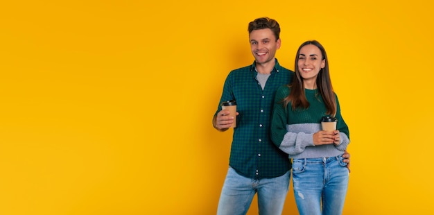 Heureux charmant jeune couple avec des tasses de café ou de thé dans les mains portant des vêtements décontractés, s'embrassant et riant ensemble tout en s'isolant sur fond jaune