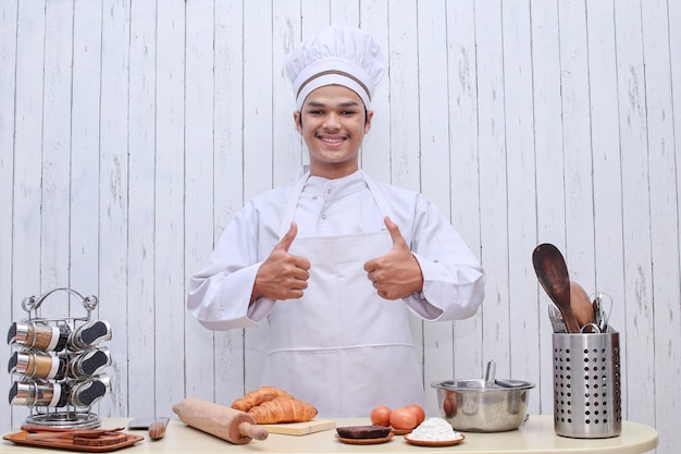 Heureux chef masculin souriant en toque montrant les pouces vers le haut dans la table de la cuisine