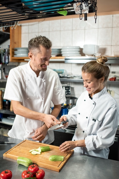 Heureux chef professionnel passant un couteau tranchant à sa stagiaire féminine coupant des courgettes fraîches tout en se tenant debout près de la table dans la cuisine du restaurant