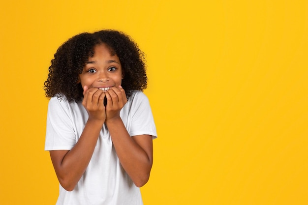 Heureux choqué jolie adolescente bouclée en t-shirt blanc mord ses doigts studio jaune isolé