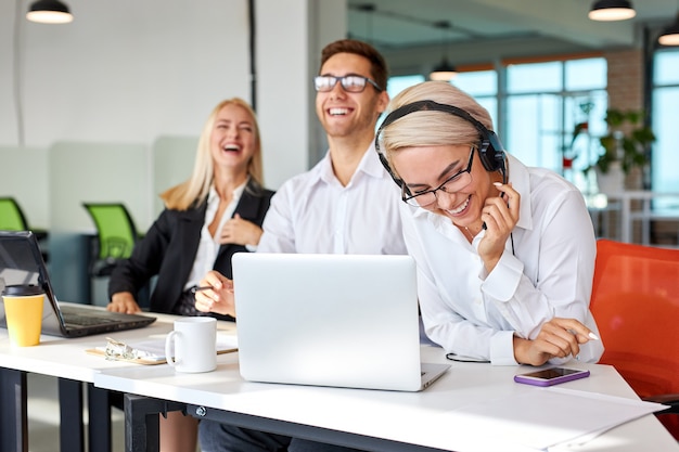 Heureux collègues riant au lieu de travail, homme et femme de race blanche s'asseoir avec un ordinateur portable s'amuser, faire une pause. se concentrer sur la femme blonde dans les écouteurs