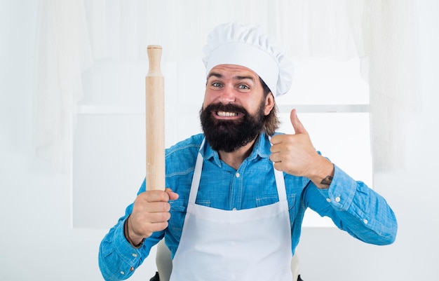 Heureux concept de cuisson de ménage homme barbu mature porter chapeau de chef tenant un rouleau à pâtisserie meilleure qualité cuisinier mâle préparer des aliments dans la cuisine hipster brutal en tablier cuisson temps de repas pour manger