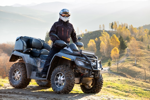 Heureux conducteur masculin dans un casque de protection profitant d'une balade extrême sur une moto quad atv dans les montagnes d'automne au coucher du soleil.