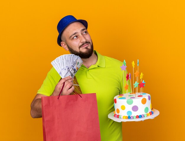 Heureux à côté d'un jeune homme portant un chapeau de fête tenant un sac-cadeau avec un gâteau et de l'argent isolé sur un mur orange