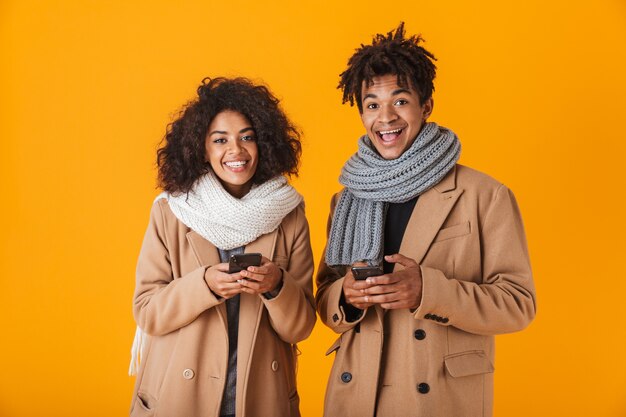 Heureux couple africain portant des vêtements d'hiver debout isolé, à l'aide de téléphones mobiles