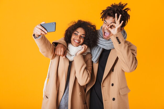 Heureux couple africain portant des vêtements d'hiver debout isolé, prenant un selfie