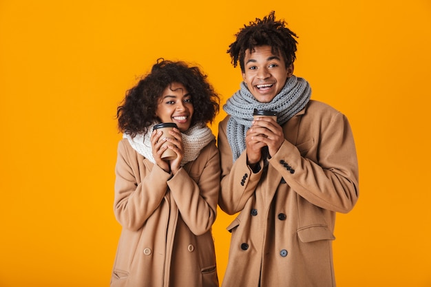 Heureux couple africain portant des vêtements d'hiver debout sous un parapluie isolé, tenant des tasses à café à emporter