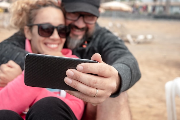 Heureux couple d'âge moyen caucasien flou assis en plein air sur la plage regardant souriant au smartphone