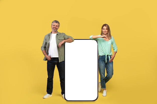 Heureux couple d'âge moyen s'appuyant sur un smartphone géant avec une maquette d'écran vierge annonçant votre service ou produit