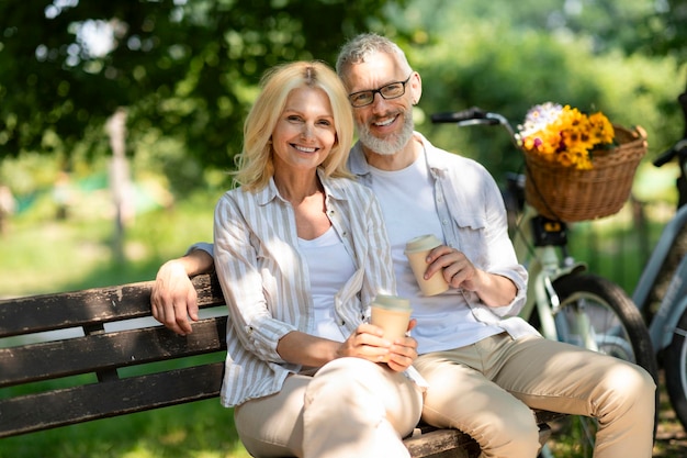 Heureux couple d'âge mûr avec café à emporter se relaxant sur un banc dans le parc