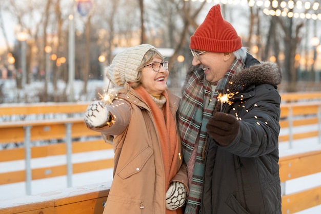 Heureux couple d'âge mûr tenant des cierges magiques et souriant les uns aux autres, ils célèbrent Noël dans le parc