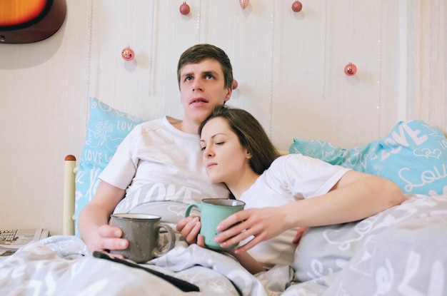 Heureux couple allongé dans son lit dans une maison confortable, buvant du café. Jeune homme en t-shirt, jolie femme. Famille