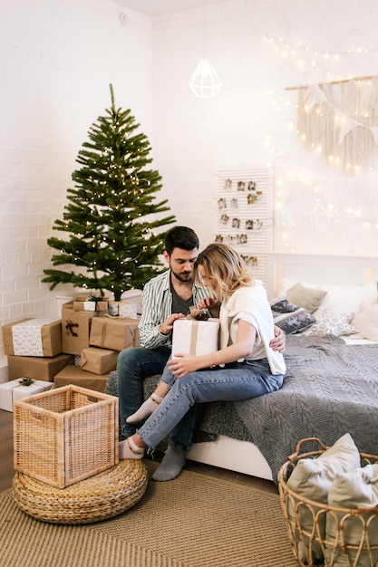 Heureux couple amoureux assis sur le lit donnant des cadeaux par l'arbre de Noël dans une maison confortable