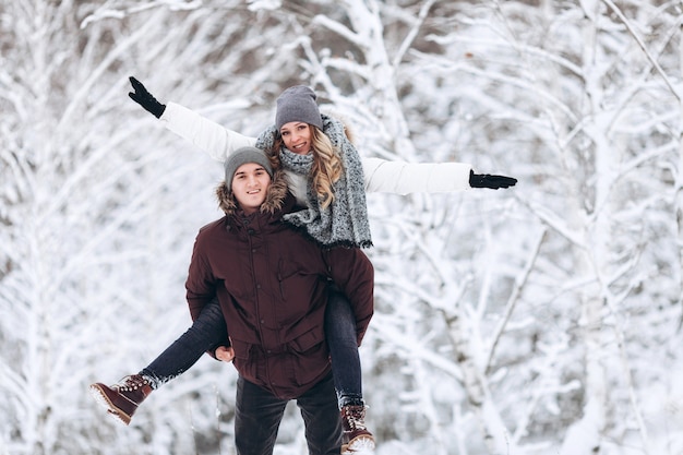 Heureux couple amoureux en hiver dans une forêt enneigée, une fille monte sur le dos de son mec bien-aimé jouant et souriant