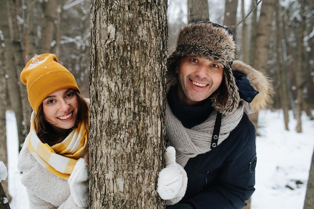 Heureux couple amoureux, ils regardent derrière un arbre, debout dans une forêt d'hiver et riant. Ils sont tous les deux habillés pour l'hiver.