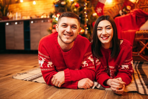 Heureux couple amoureux se trouve sur le sol, vacances de Noël. Célébration de Noël, joyeux homme et femme en pulls rouges, sapin avec décoration