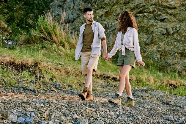 Heureux couple amoureux en tenue décontractée se regardant et discutant de quelque chose tout en se tenant par les mains et en descendant la berge