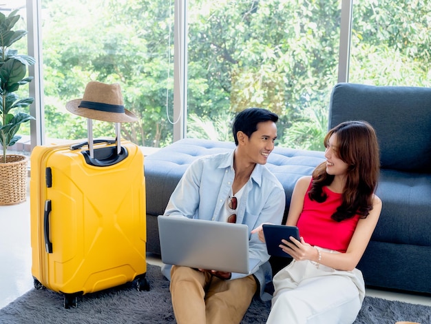 Heureux couple asiatique jeune homme et femme se regardant tout en utilisant un ordinateur portable et une tablette ensemble pour la réservation de vol et les informations de voyage dans le salon près des bagages vacances d'été